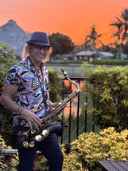 Smiling woman in a colorful geometric shirt and blue fedora playing a saxophone outdoors at sunset near greenery.