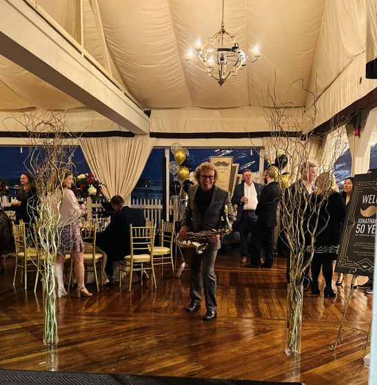 Male saxophonist playing a brass saxophone at a formal indoor party with guests and gold chairs in the background.