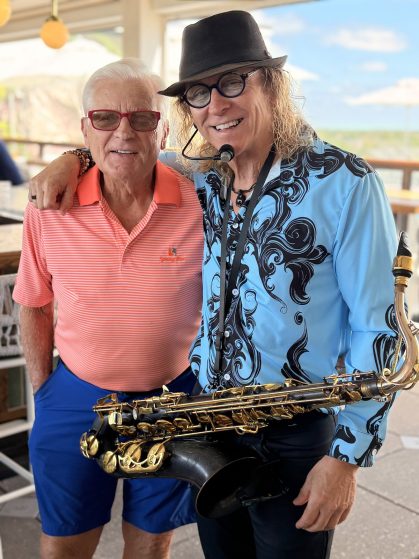 Two smiling men posing outdoors, one in an orange striped polo and red glasses, the other in a black hat with a blue patterned shirt, holding a saxophone between them.