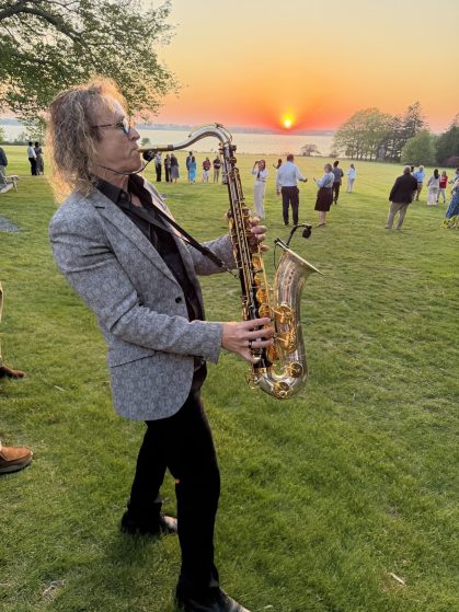 Saxophonist in a gray patterned blazer playing outdoors at sunset, with guests mingling on a lawn by a lake in the background.