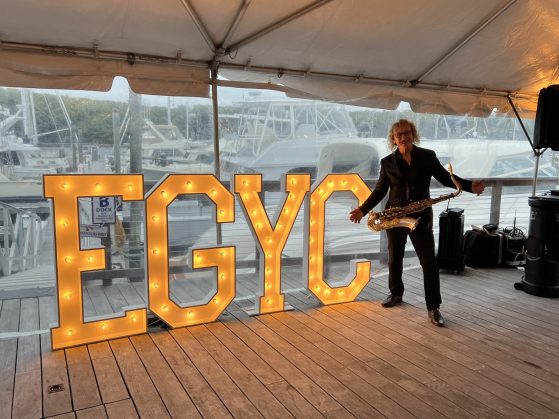 Saxophonist in a black suit stands beside oversized illuminated letters spelling 'EGYC' on a wooden marina deck.