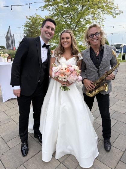 MattyB posing with the bride and groom holding his saxophone at an outdoor waterfront wedding venue