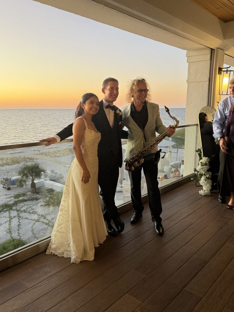 MattyB holding his saxophone with the bride and groom on a balcony overlooking the beach at sunset