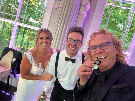 MattyB posing with the bride and groom in front of tall windows at an elegant purple-lit wedding reception
