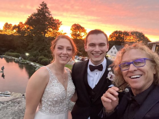 MattyB posing with the bride and groom in front of a dramatic sunset over the water