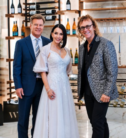 MattyB posing with a couple in formal attire in front of a wine cellar display at a wedding celebration
