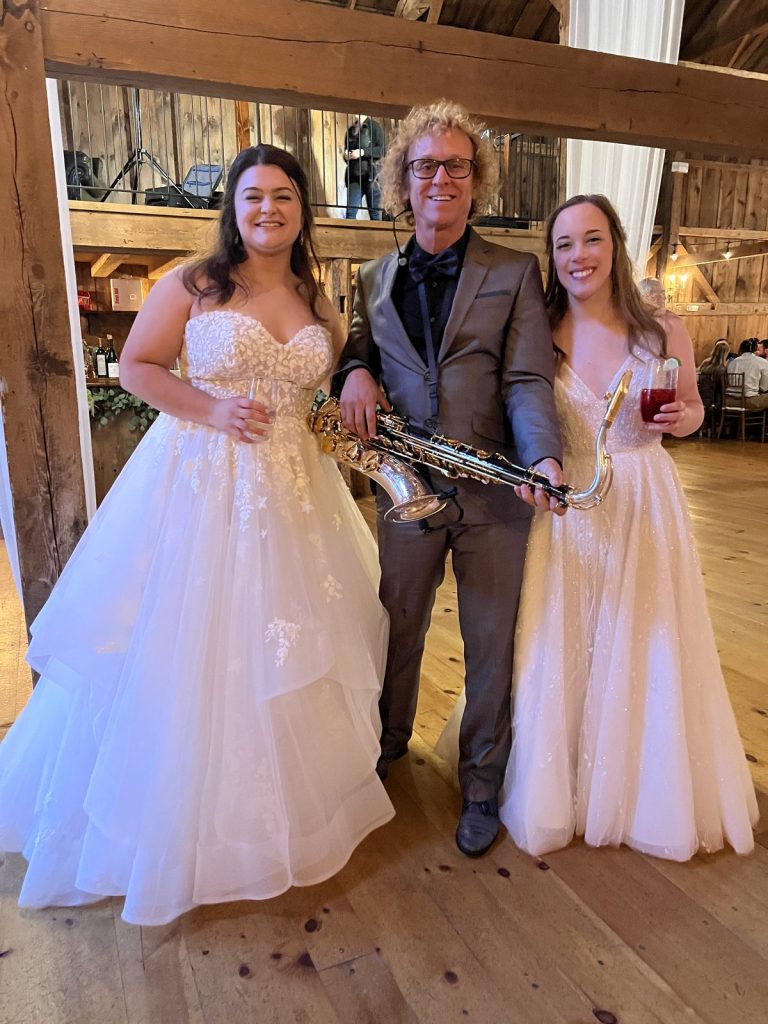 MattyB holding his saxophone out between the bride and a bridesmaid in white dresses at a rustic barn wedding venue