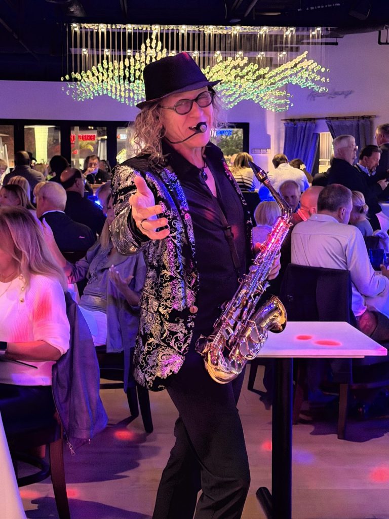 MattyB performing on alto saxophone with a headset mic among guests at a colourfully lit Southwest Florida venue.