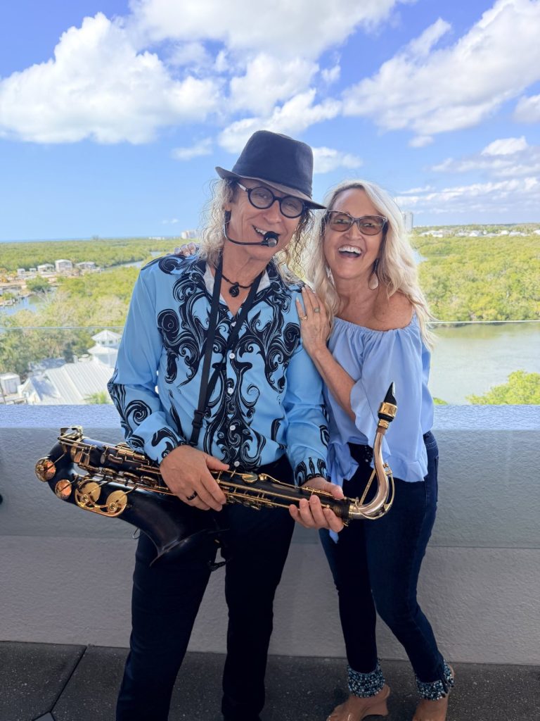 MattyB in a patterned blue stage shirt and black hat holding his saxophone, laughing with a smiling blonde woman at a waterfront venue in Southwest Florida.