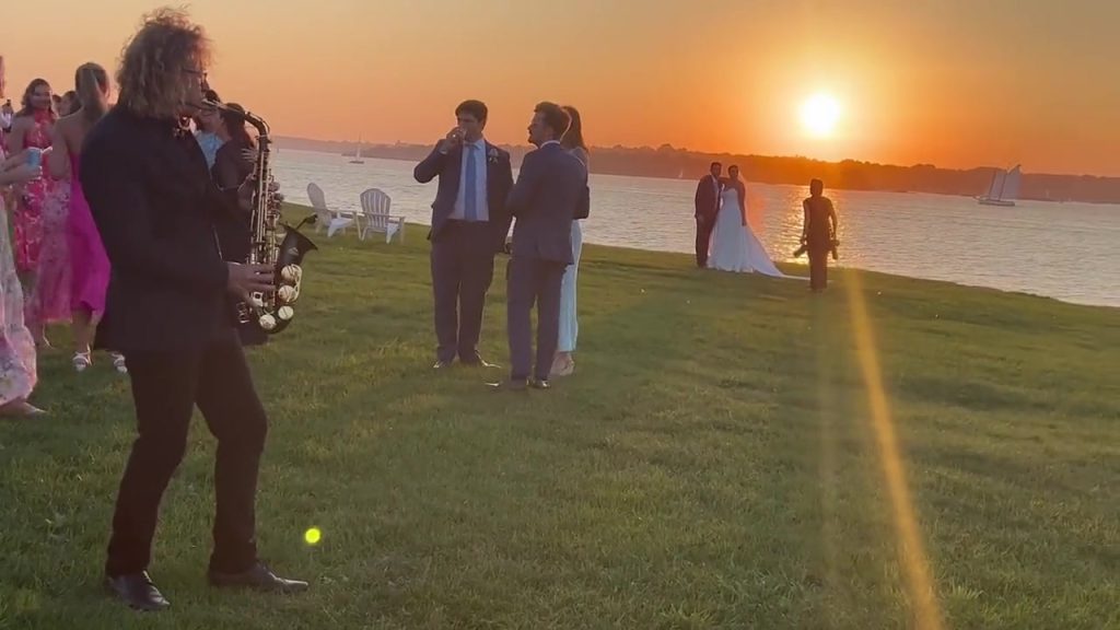 MattyB playing saxophone at golden hour as a couple watches the sunset during wedding cocktail hour
